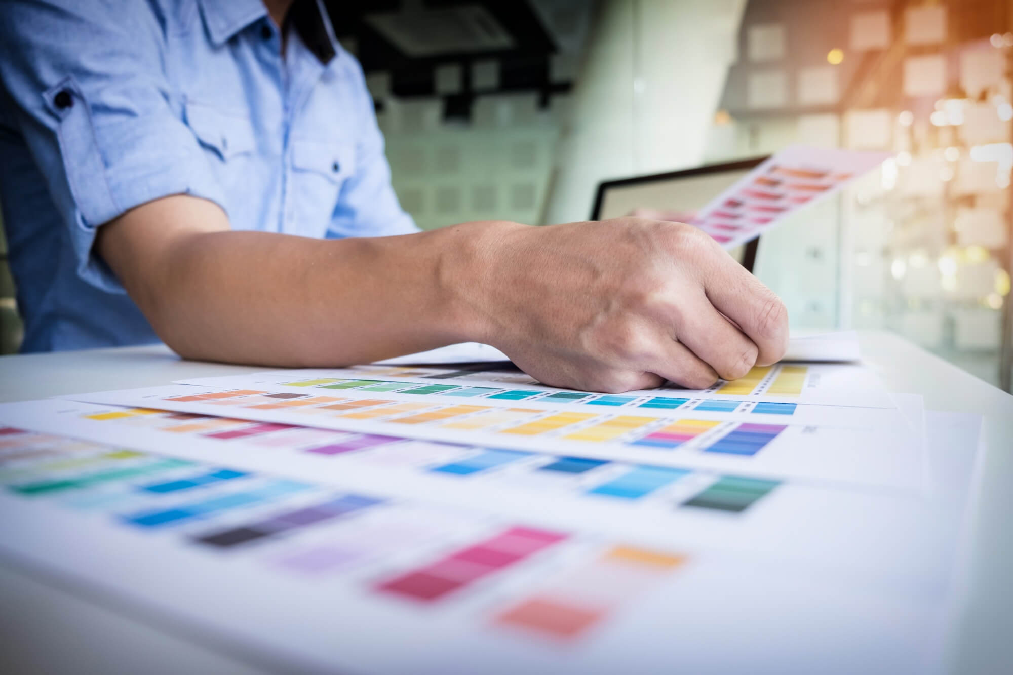 Diseñador gráfico utilizando una tableta digital y guías de color Pantone en un escritorio de madera con notas adhesivas y una taza de café.